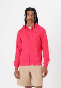Roza zip-up kapuca s logotipom "GAP", s kapuco na vezanje, kenguru žepima i rebrastim manžetama, u kombinaciji s bež kratkim hlačama.