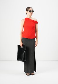 Mujer que lleva gafas de sol negras, top rojo de un solo hombro, falda larga negra con abertura lateral, sostiene un bolso tote de cuero negro y lleva sandalias.