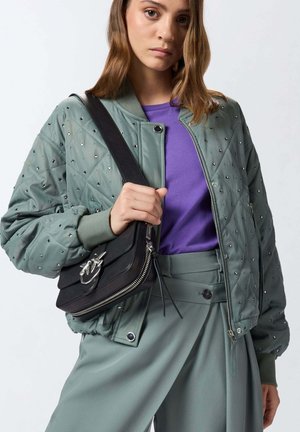 Giovane donna che indossa una giacca trapuntata verde con borchie, una camicia viola, pantaloni verdi e tiene una borsa a tracolla in pelle nera.