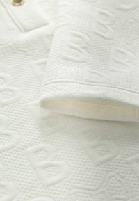 Tela blanca texturizada con patrones en relieve de "B" por todo. Presenta un puño doblado y un botón en el escote. Textura suave y tejida.