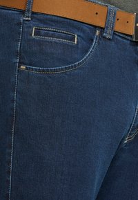 Donkerblauwe denim jeans met gele stiksels, voorzak en een bruine leren riem met een zilveren gesp.