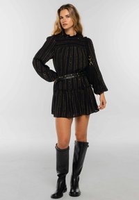 Vestido negro con rayas verticales doradas, dobladillo con volantes y mangas largas; combinado con botas negras altas hasta la rodilla y un bolso pequeño negro.