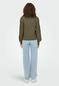 Maglione verde oliva con texture, dal taglio ampio, orlo arrotondato e maniche a sbuffo; abbinato a pantaloni in denim azzurro dritti e sneakers bianche.