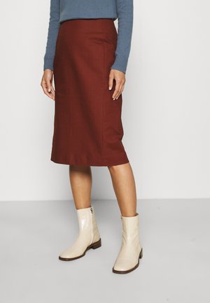 Person iført et rustfarvet knælange nederdel, lyse ankelstøvler og en blå langærmet sweater, stående på et gråt gulv.