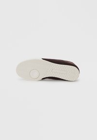 Sapato slip-on masculino em camurça castanha com sola texturizada branca, apresentando padrões de aderência circulares e em zig-zag.