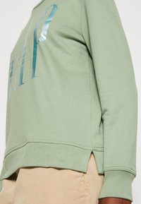 Felpa verde chiaro con un logo blu lucido, vestibilità rilassata, spacchi laterali e texture morbida. Presenta polsini e orlo a costine.