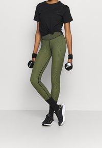 Camiseta negra con nudo en el frente, leggings a rayas verdes, zapatillas deportivas negras y muñequeras. Sostiene mancuernas.