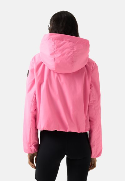 Person von hinten zu sehen, trägt eine leuchtend pinke Kapuzenjacke und schwarze Leggings vor weißem Hintergrund.