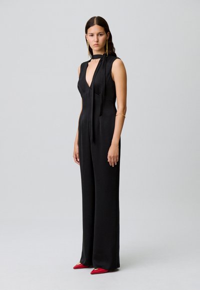 Mono negro sin mangas con un profundo escote en V y un detalle de lazo en el cuello, con patas anchas y una textura suave. Combinado con tacones rojos de punta.