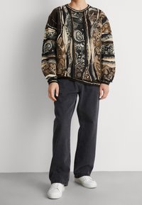 Sweter dziany z wielokolorowym geometrycznym wzorem, w odcieniach czerni, brązu i kremu. Połączony z ciemnoszarymi spodniami i białymi butami.