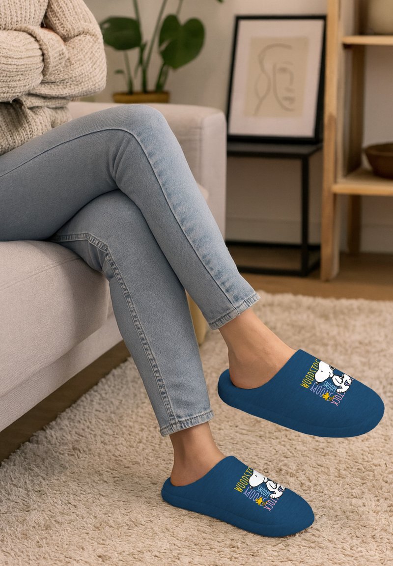 Pantofole blu senza lacci con una texture morbida, caratterizzate da una grafica di cartone animato e testo colorato sulla parte superiore. Indossate con jeans blu chiaro.
