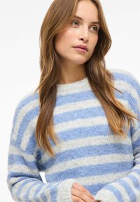 Mujer joven con cabello largo castaño claro, vistiendo un suéter peludo a rayas azul y blanco, mirando ligeramente de lado.