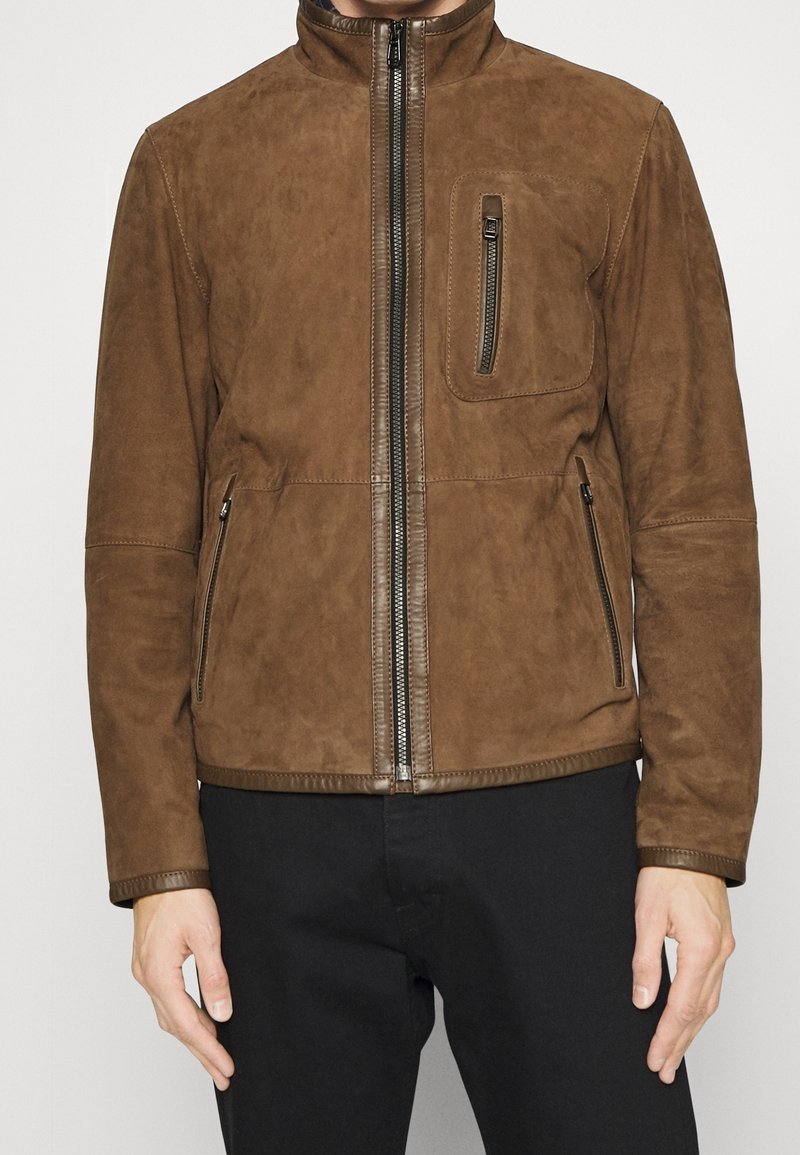 Brun mockaskinnjacka med hög krage, frontdragkedja och bröstficka. Har sidofickor och ribbade ärmslut, vilket tillför struktur och textur.