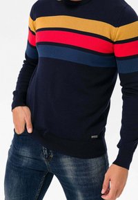 Pull marine à rayures horizontales rouges, jaunes, bleu foncé et bleu clair. Encolure et poignets côtelés. Associé à un jean en denim foncé.