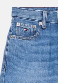 Lyseblå denimjeans med en frontlomme, metallnitter og en liten logo-patch med røde, hvite og blå farger på lommen.