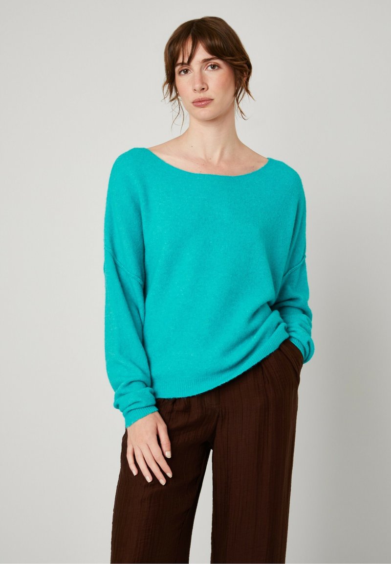 Turkoois oversized sweater met een wijde halslijn en lange mouwen, gecombineerd met getextureerde bruine broeken. Zachte, gebreide stof.