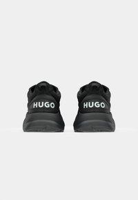 Černé sportovní boty s aerodynamickým designem, vybavené strukturovaným svrškem, silnou podešví a logem "HUGO" na patě.