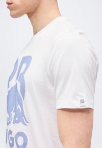 Νεαρός άνδρας με λευκή κοντομάνικη μπλούζα με μεγάλο γαλάζιο γραφικό και μικρό λογότυπο "HUGO" στο μανίκι, απεικονιζόμενος από το πλάι και από το λαιμό και κάτω.