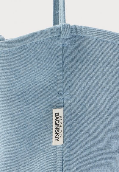 Γωνία τσάντας tote από ανοιχτό μπλε τζιν με λεπτομέρεια ραφής και λευκή υφασμάτινη ετικέτα που γράφει "Ruslan Baginskiy", προσαρτημένη κοντά στη ραφή.