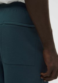 Pantalones jogger en color azul verdoso con una textura suave, que cuentan con un bolsillo trasero rectangular y una cintura elástica. Primer plano mostrando una mano sobre la tela.