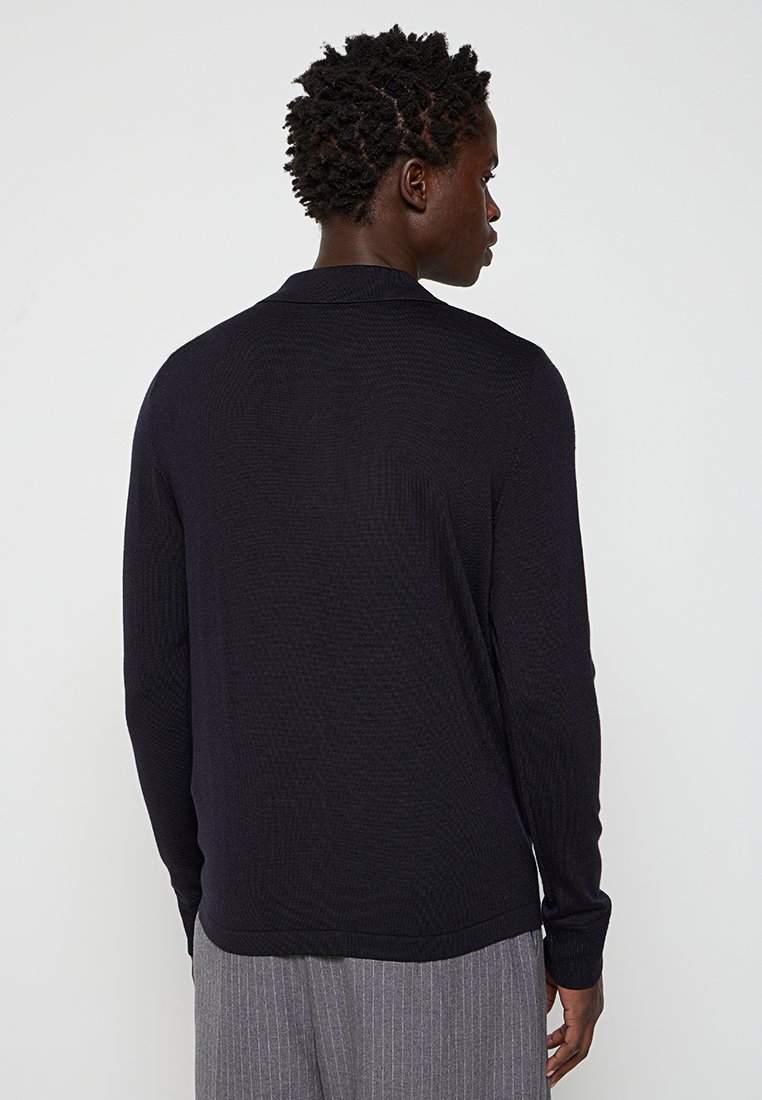 Suéter de punto negro con un acabado texturizado, mangas largas y un cuello. Presenta un corte recto y un minimalismo en los detalles en la parte trasera.
