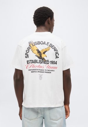 Man facing away wearing a white T-shirt with a large yellow eagle and black and red text referencing Sport Lisboa e Benfica and "Established 1904."