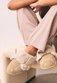 Chaussons beiges doux en tissu texturé, avec des cornes décoratives et des accents verts. Doublure en fourrure douce et détail en corde cousue sur la semelle.