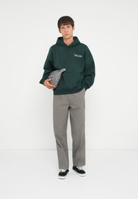 Dunkelgrüner Kapuzenpullover mit weißem Logo, graue Bauchtasche, graue und weiße gemusterte Hosen und schwarze Sneakers mit weißen Sohlen.