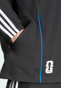 Sort atletisk jakke med hvide og grønne striber, lynlåslomme, blå detaljesyninger og et hvidt FIFA-logo nær kanten.