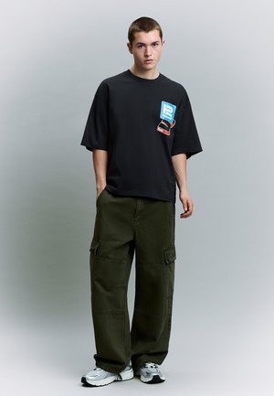 Jeune homme debout portant un t-shirt graphique noir, un pantalon cargo vert olive et des baskets blanches de sport sur un fond gris.
