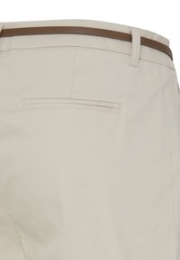 Un pantalon beige avec une seule poche passepoilée au dos et une fine ceinture marron passée dans les passants.