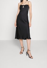 Černé saténové midi šaty s tenkými ramínky, detaily ve tvaru klíčové dírky a mašlí u výstřihu. Hladká textura a splývavý silueta, doplněné černými lodičkami.