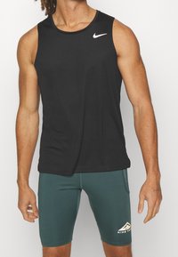 Personne athlétique portant un débardeur noir Nike et un short Nike Trail vert, debout devant un fond blanc.