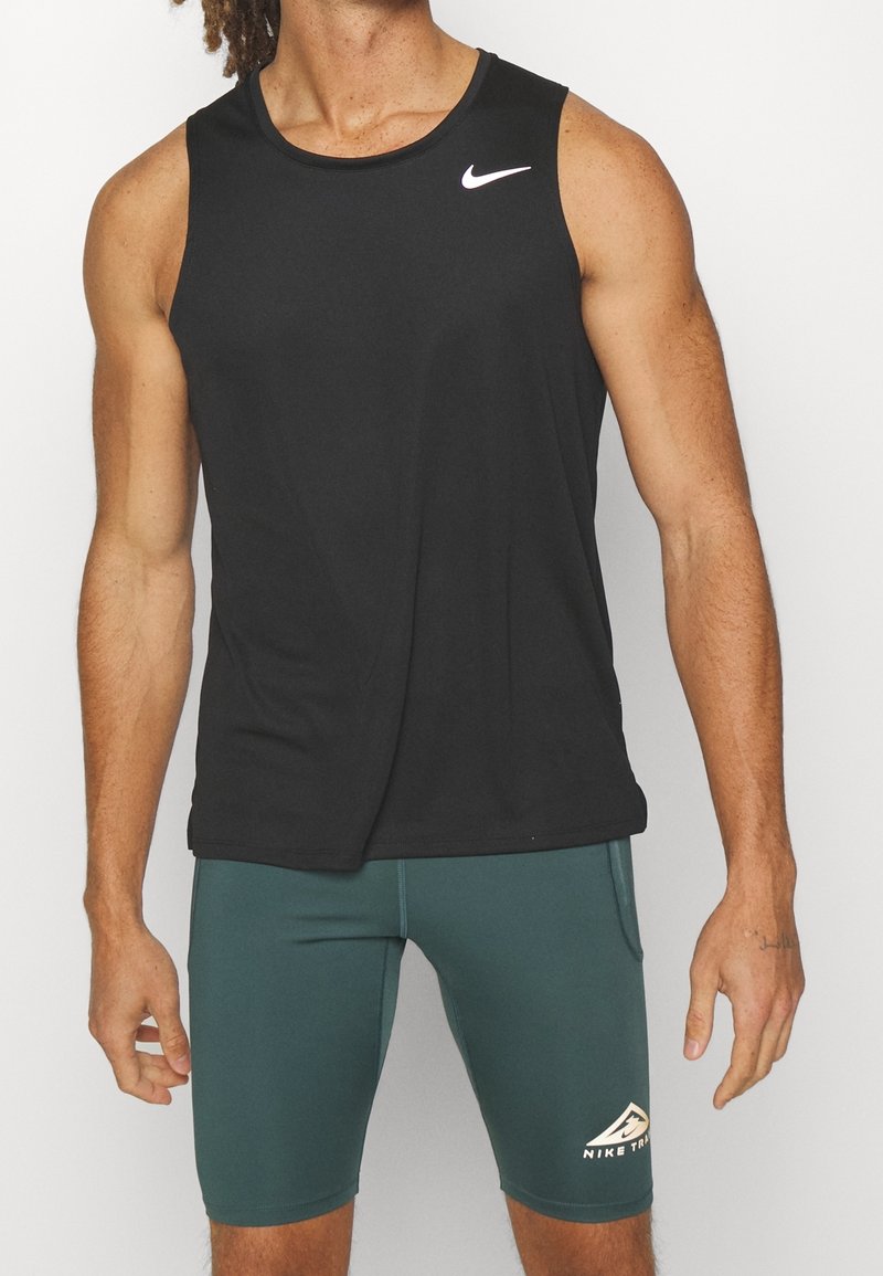 Personne athlétique portant un débardeur noir Nike et un short Nike Trail vert, debout devant un fond blanc.