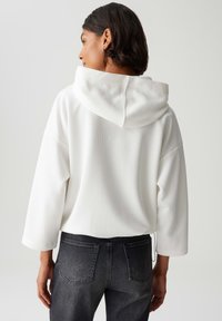 Vrouw met kort donker haar, gekleed in een textuurrijke witte hoodie en zwarte jeans, gezien vanaf de achterkant tegen een effen lichte achtergrond.
