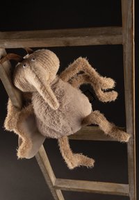 Plush toy resembling a creature with a round body, long beak, beige and khaki fur, and flexible limbs, positioned on a wooden ladder.