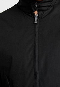 Närbild av en svart jacka med dragkedja framtill och två svarta knappar vid kragen, som visar tygtextur och sömnadsdetaljer.