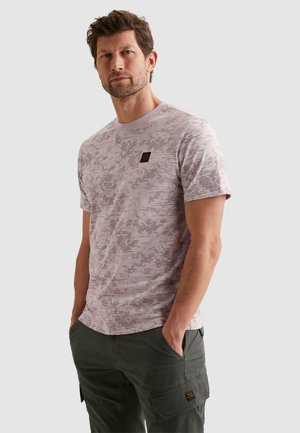 Muž s krátkymi vlasmi a bradou, oblečený v svetlom tričku s potlačou a tmavých cargo nohaviciach, ruky v kapse, neutrálny pozadie.