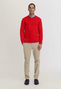 Roter Strickpullover mit V-Ausschnitt, kombiniert mit einem hellblauen Hemd mit Kragen, khakifarbenen Hosen und schwarzen Schuhen. Einfaches Design, lässige Passform.