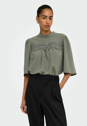 Femme avec queue de cheval portant un chemisier vert avec un détail en dentelle et un pantalon noir taille haute, debout avec une main dans la poche sur un fond clair.
