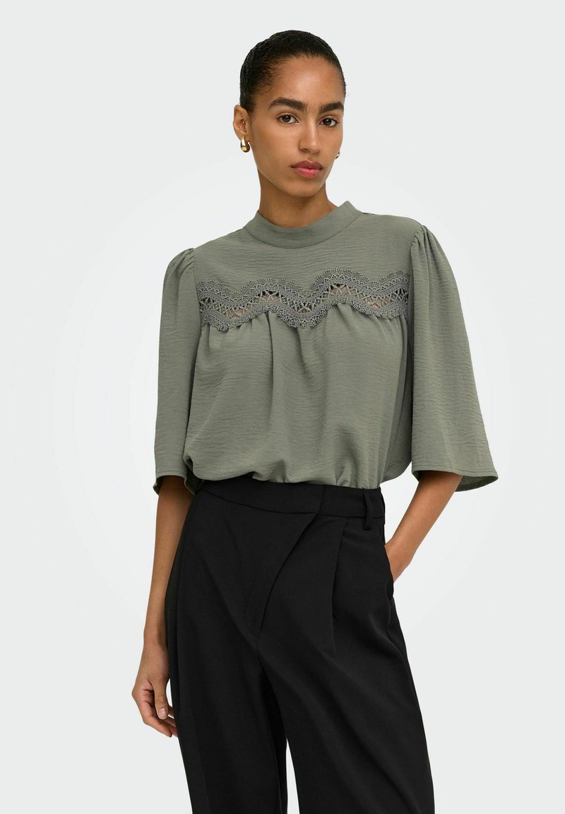 Femme avec queue de cheval portant un chemisier vert avec un détail en dentelle et un pantalon noir taille haute, debout avec une main dans la poche sur un fond clair.