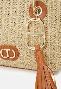 Käsin kudottu olkalaukku tehty oljesta beigen värisenä, jossa on ruskeat nahkakoristeet. Varustettu kullansävyisellä sulkijalla ja ruskealla nahkatupsu yksityiskohdalla.