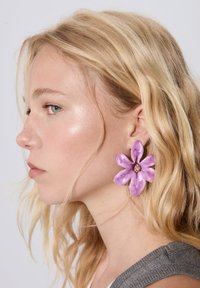 Veľká fialová náušnica v tvare kvetu, vyrobená z lesklého materiálu, s centrálnym drahokamom. Nosená na modelke so svetlou pokožkou a vlnitými blonďavými vlasmi.