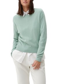 Maglione verde chiaro con un motivo di lavorazione a maglia testurizzata, polsini e vita a coste, indossato sopra una camicia bianca con colletto.