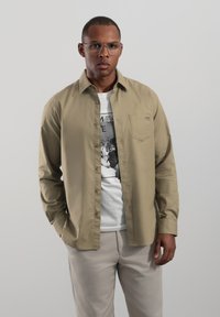 Chemise kaki clair à manches longues avec un design à boutons, une poche poitrine unique et une texture lisse, portée par-dessus un t-shirt blanc à motif.