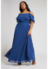 Blå maxi-klänning med lager av volangdetaljer på axlarna, elastisk midja, flödande material och ankellång kjol. Klara snörskor.