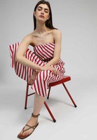 Femme assise sur une chaise rouge, portant une combinaison sans bretelles à rayures rouges et blanches et des sandales à lanières marron, sur un fond uni.