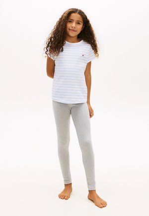 Νεαρή κοπέλα που στέκεται ξυπόλυτη, φορώντας ένα ανοιχτό μπλε ριγέ μπλουζάκι και γκρι κολάν, μπροστά σε λευκό απλό φόντο.