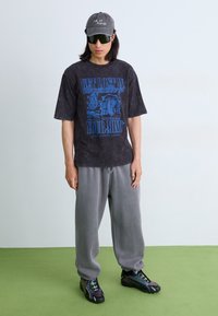 Overdimensioneret sort graphic t-shirt med blå tekst, grå sweatpants og sorte sko med grønne accenter, suppleret med en grå kasket og solbriller.