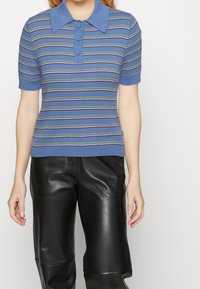 Blå kortärmad polo tröja med krage, prydd med horisontella ränder i grönt och vitt. Bärs tillsammans med svarta läderbyxor.